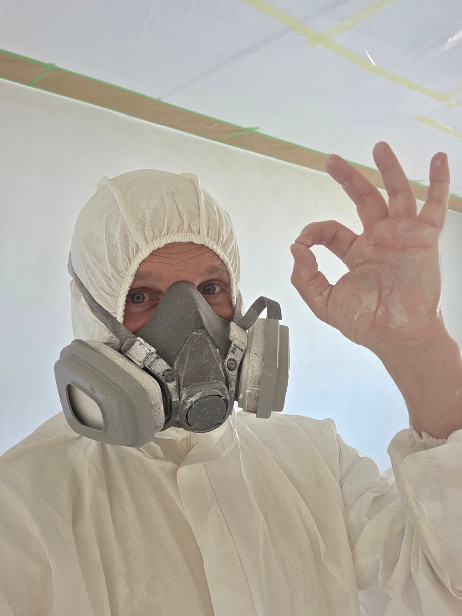 Stukadoor in beschermpak met masker steekt duim en vinger op tijdens het spuitwerk