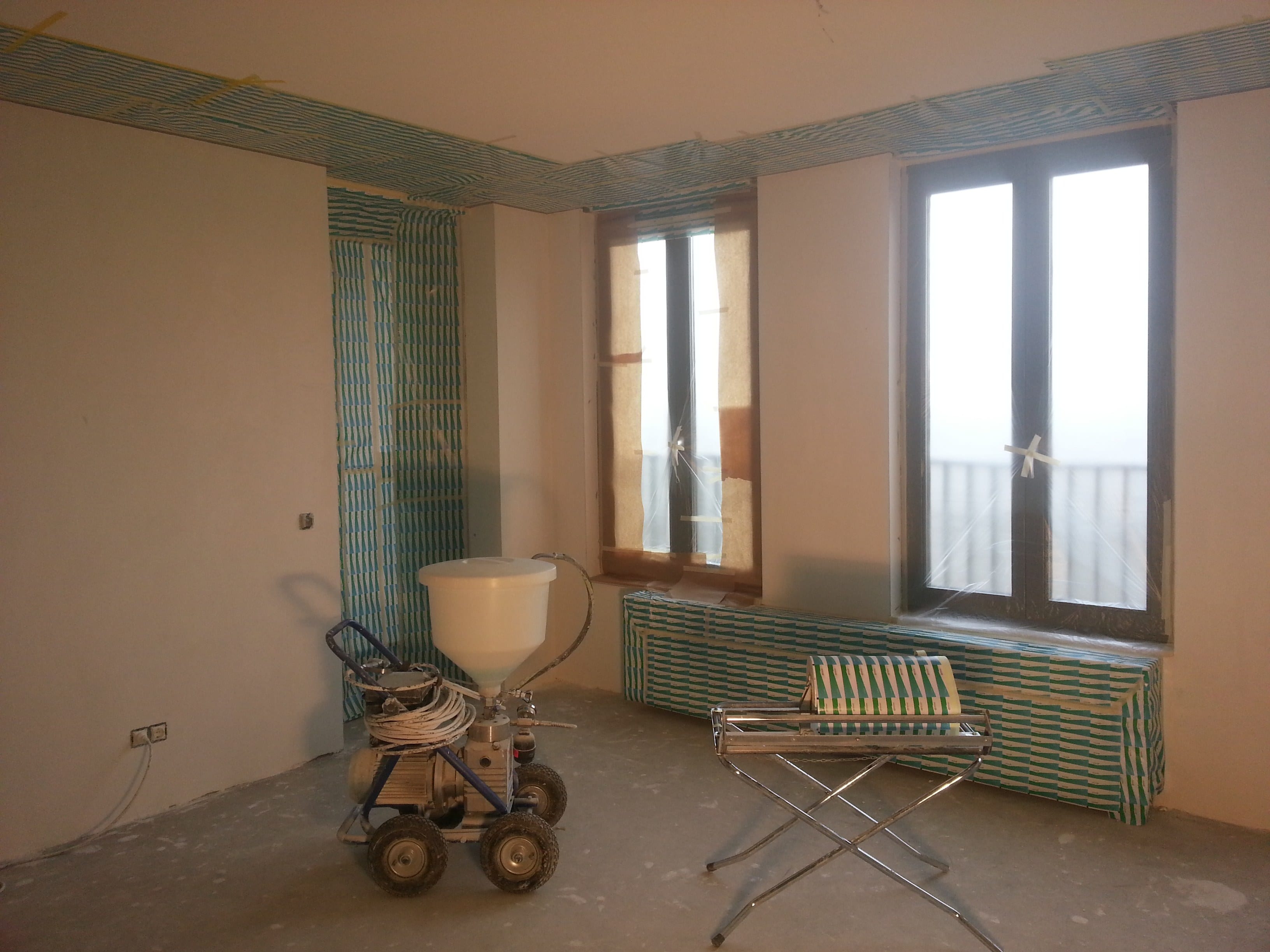 Kamer volledig afgeplakt rond kozijnen en plafondrand, met de spuitinstallatie klaar voor gebruik