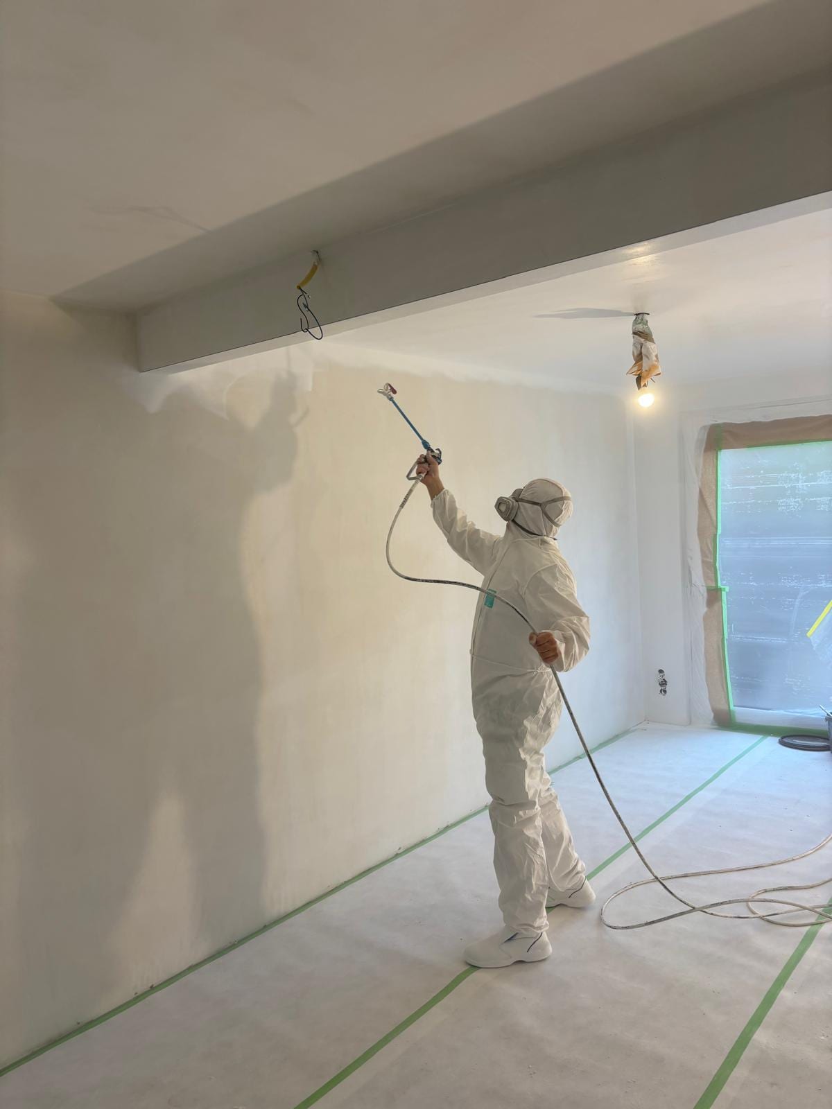 Stukadoor spuit van opzij gezien de wand van een afgeplakte ruimte, spuitpistool schuin omhoog