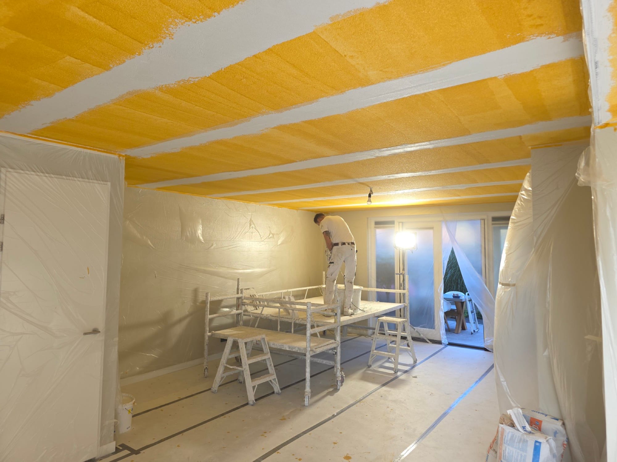 Stukadoor aan het werk in een afgeplakte ruimte, het plafond in gele voorstrijk met witte banen